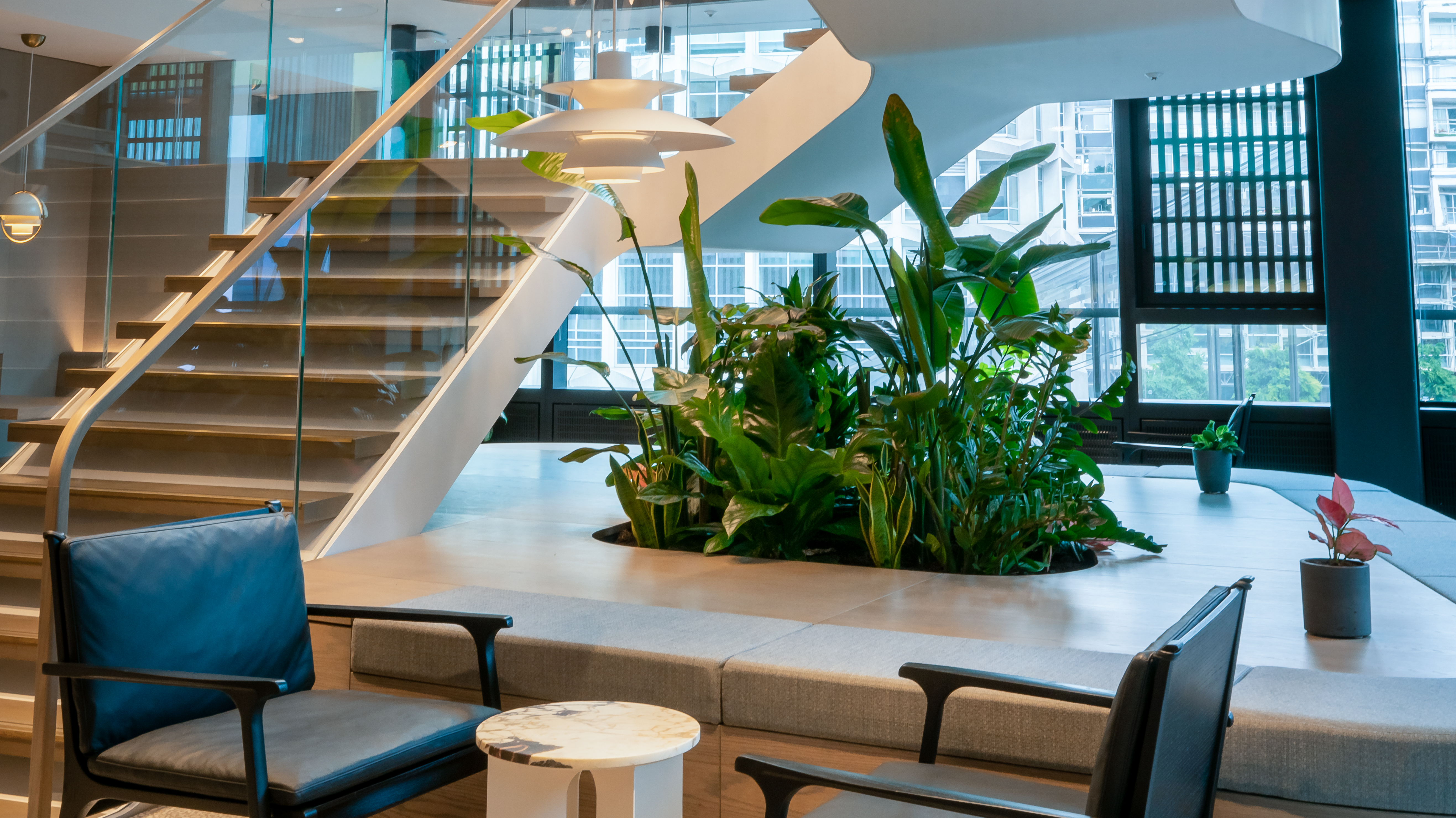Modern office lounge with indoor plants, glass stairway, natural lighting, and contemporary seating, reflecting a professional and sustainable work environment.