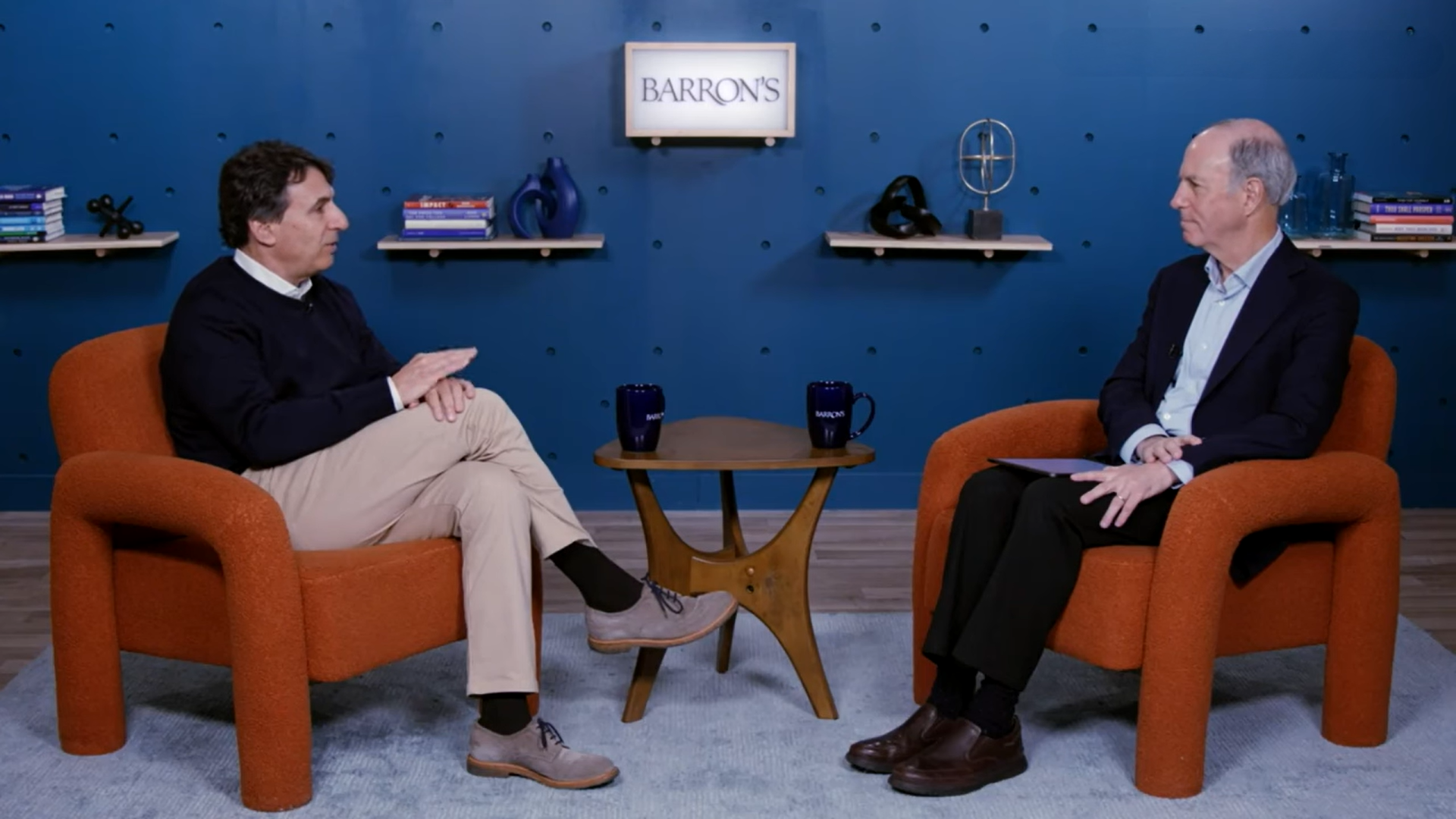 Marc Rowan and host seated in orange chairs during Barron’s interview in a blue-walled studio, discussing private markets, alternative investments, and capital solutions.