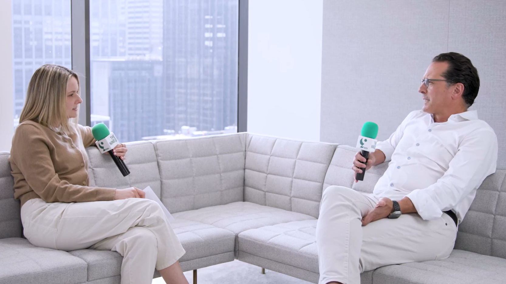 Two Apollo employees seated on a modern gray sofa in a high-rise office, engaged in a casual conversation with microphones during a 9@9 episode.