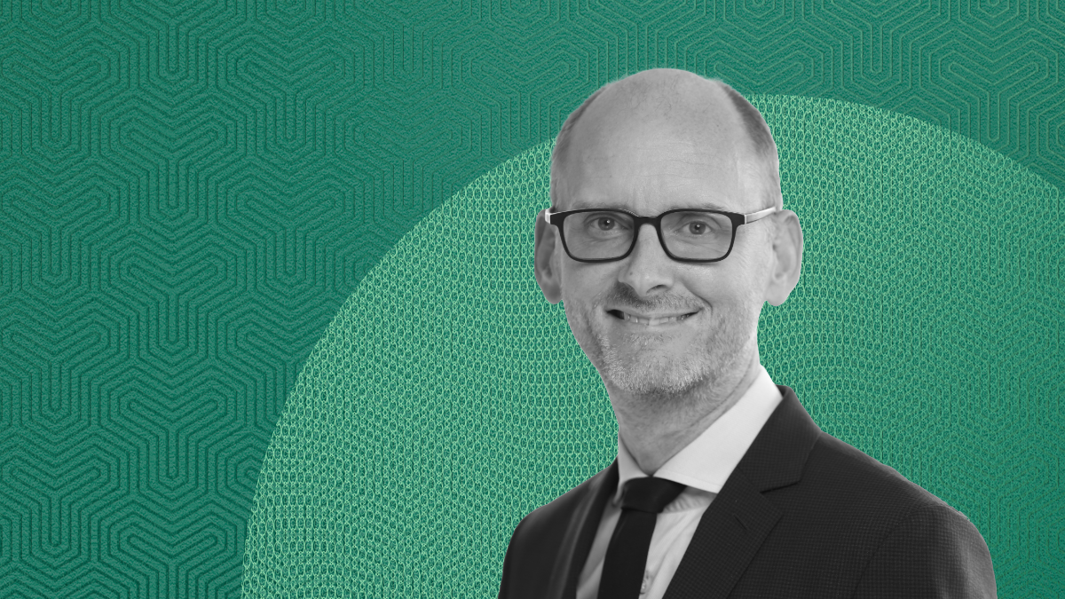Black-and-white portrait of Torsten Slok in glasses and a suit, smiling in front of a teal geometric background, discussing US economic outlook.